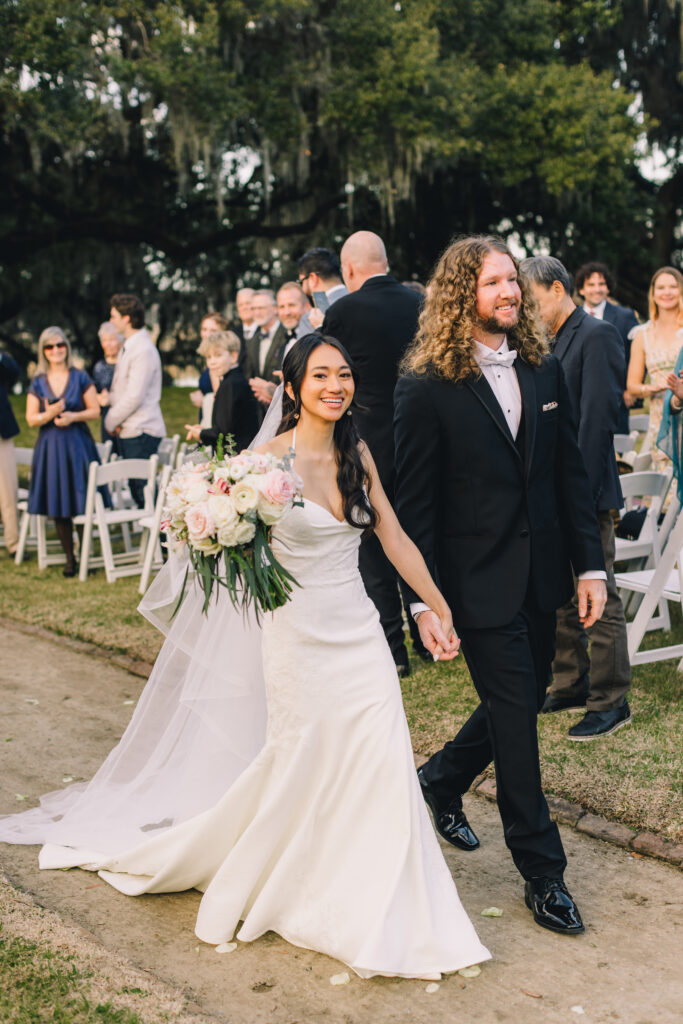 A bride beams with joy while holding a bouquet of pastel flowers, walking hand in hand with her groom as guests look on, smiling and clapping.