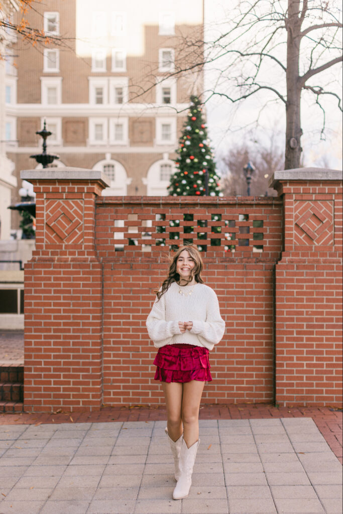 Teen girl in a white sweater, red velvet ruffle skirt, and white boots standing in front of a brick wall with a decorated Christmas tree behind her in easy photo spots in Greenville SC