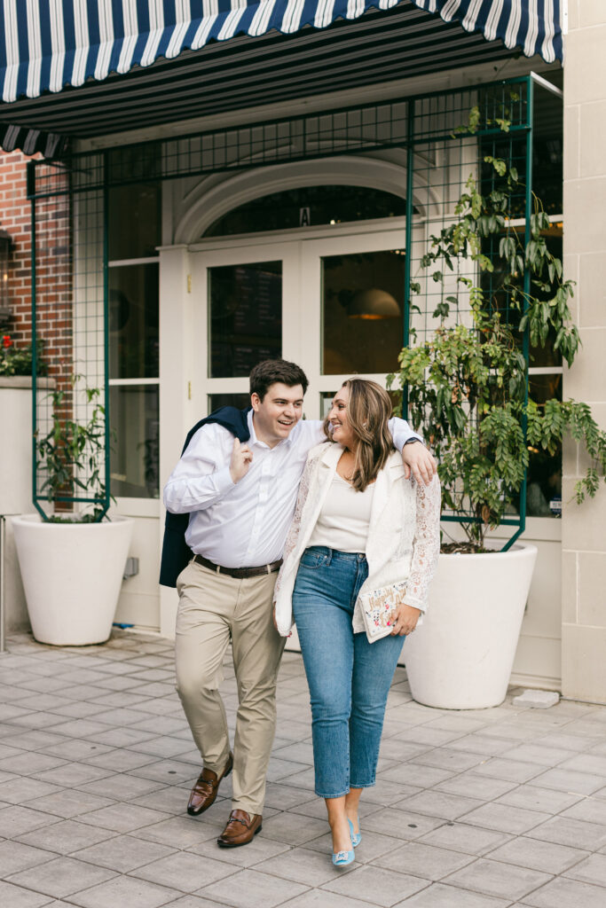 Engaged couple walking arm-in-arm in front of a striped awning downtown Greenville SC; the woman wears blue heels and a lace blazer. Dani Lacey Photographs