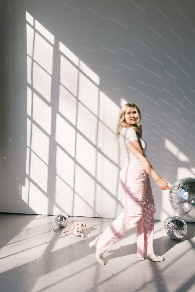Playful brand photography of a blonde woman in pink pants holding a disco ball in dramatic window light. Captured in Greenville SC by Dani Lacey Photographs.