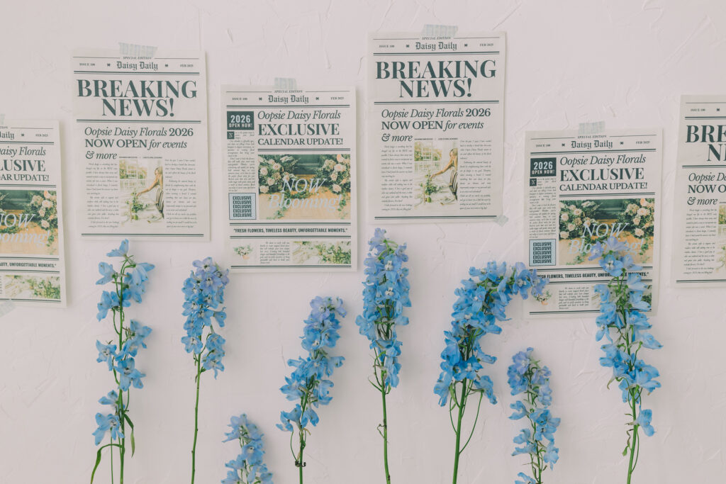 Creative flat lay of floral announcement posters and blue delphinium stems on a white textured wall — a bold reminder that it’s time for a visual rebrand, styled by Greenville South Carolina branding photographer Dani Lacey.