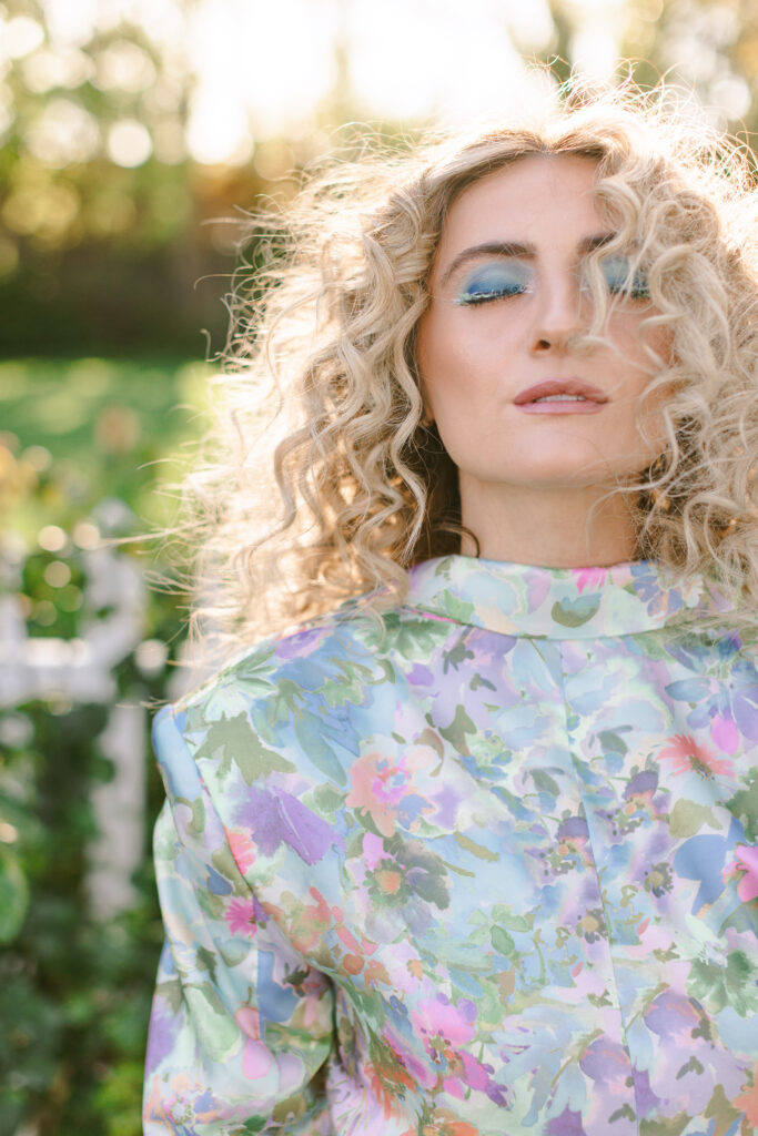 Editorial beauty brand headshot of a woman with curly blonde hair and vibrant blue eyeshadow in a floral outfit, taken outdoors in soft natural light – Greenville South Carolina Branding Photographer, Dani Lacey Photographs.