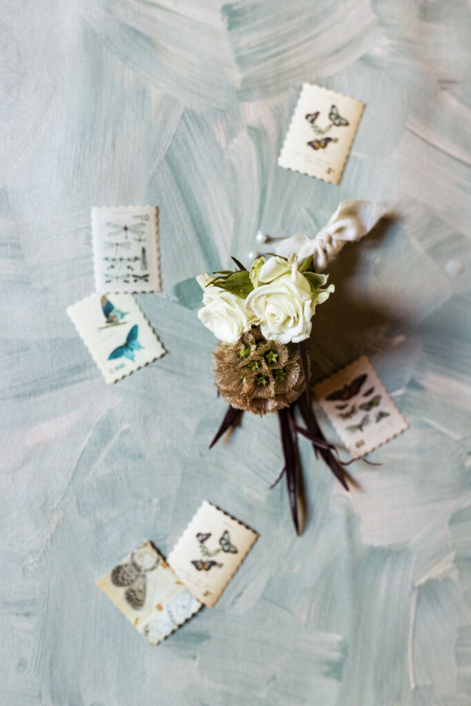 A flat lay of a groom’s boutonniere surrounded by vintage postage stamps, styled against a painterly blue background for Wedding Timelines That Work