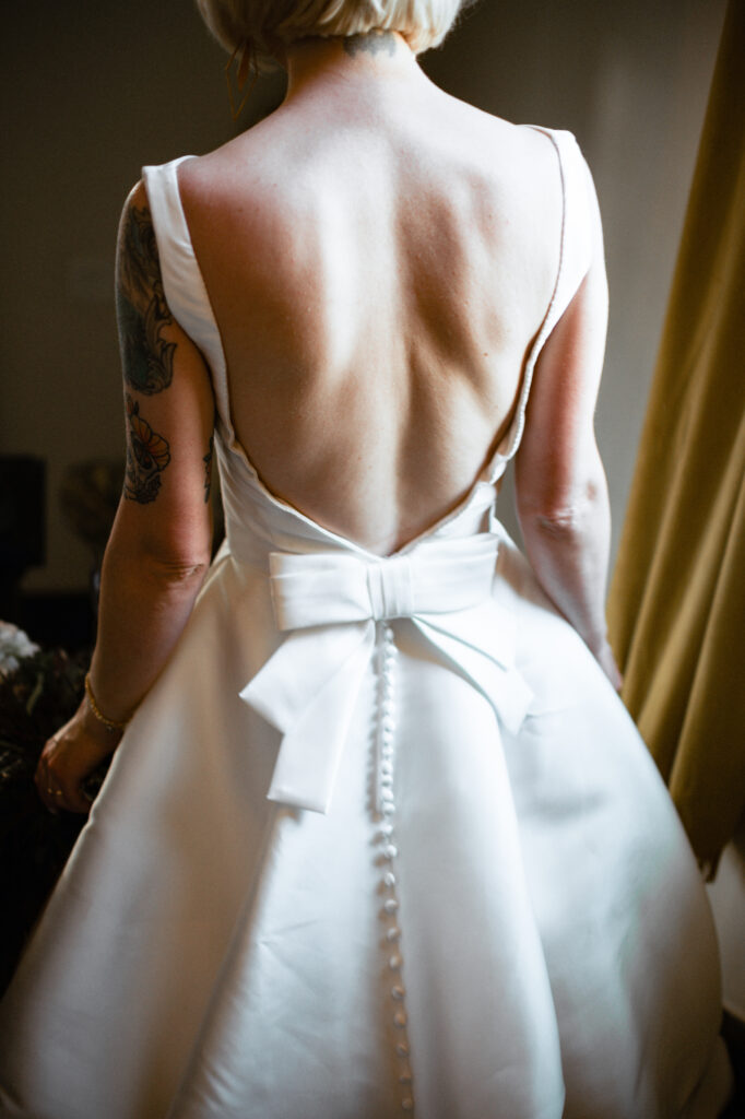 A bride stands in soft natural light, showcasing the back of her dress before the ceremony — a quiet moment that deserves space in every wedding photo timeline. Dani Lacey Photographs.