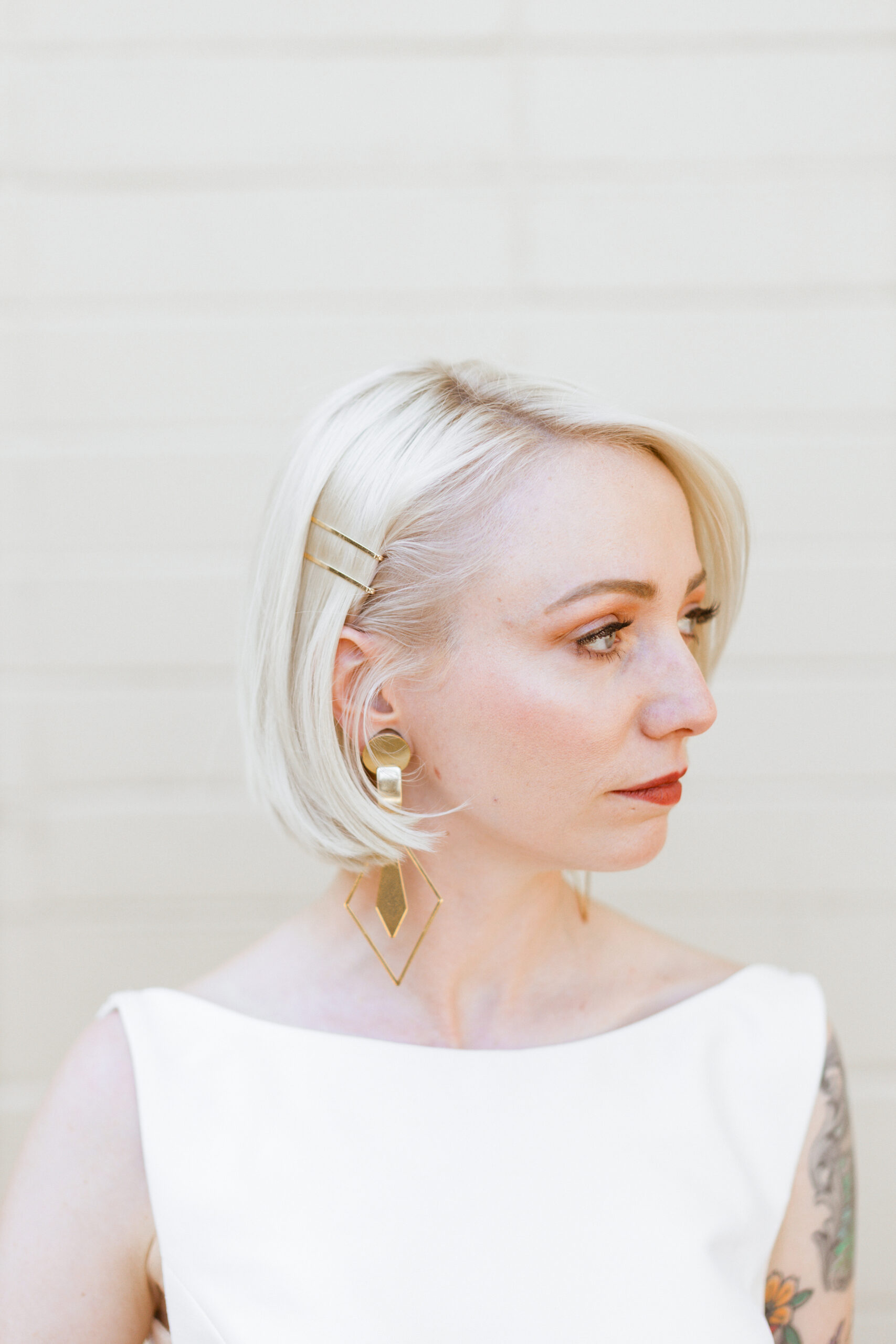 A bride with platinum blonde hair and modern gold earrings gazes softly to the side, wearing a white dress in front of a cream wall for building a wedding timeline that works