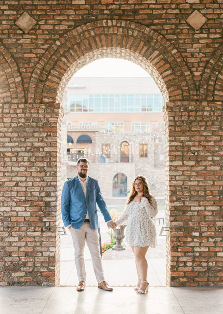 Couple standing beneath a tall brick archway at Bridgeway Station in Greenville, South Carolina, holding hands during their engagement session – Dani Lacey Photographs.