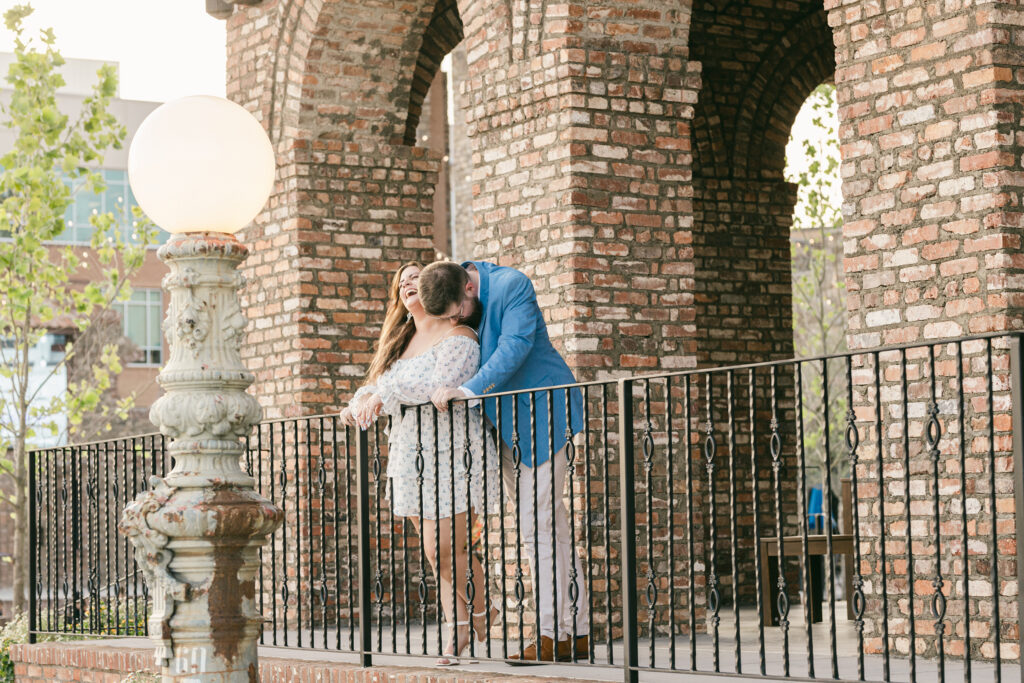 Fiancé kisses his partner's shoulder as she laughs on the iron balcony at Bridgeway Station in Greenville SC – Dani Lacey Photographs.