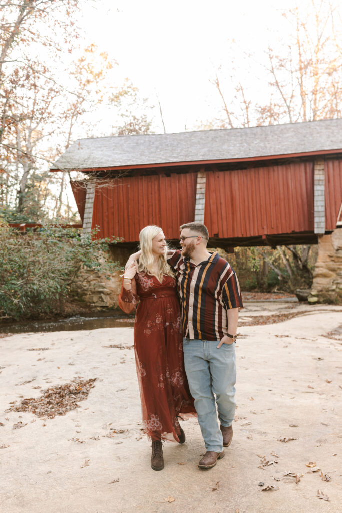 Couple walking under red covered bridge arm in arm during fall engagement shoot in South Carolina. Dani Lacey Photographs.