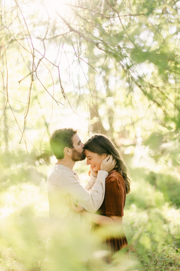 Couple sharing an intimate forehead kiss under glowing trees at Clemson Botanical Gardens during golden hour
