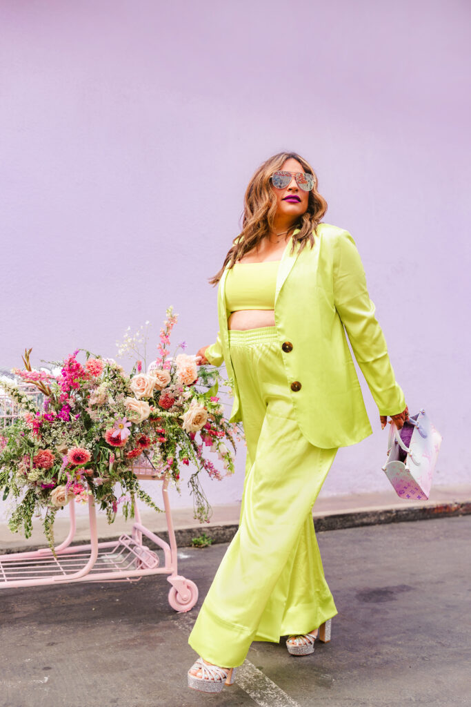 A confident woman in a chartreuse suit walks past a pastel wall and overflowing flower cart, captured during a vibrant editorial branding session by Greenville South Carolina Branding Photographer Dani Lacey.