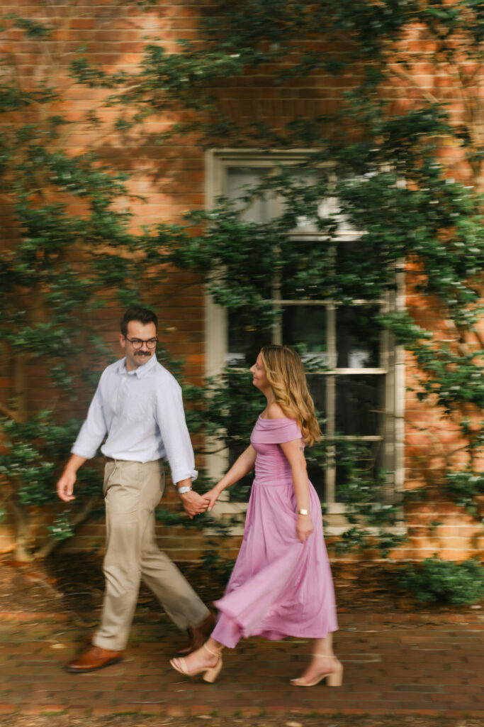 A playful couple walks past a red brick wall with ivy at Furman University.