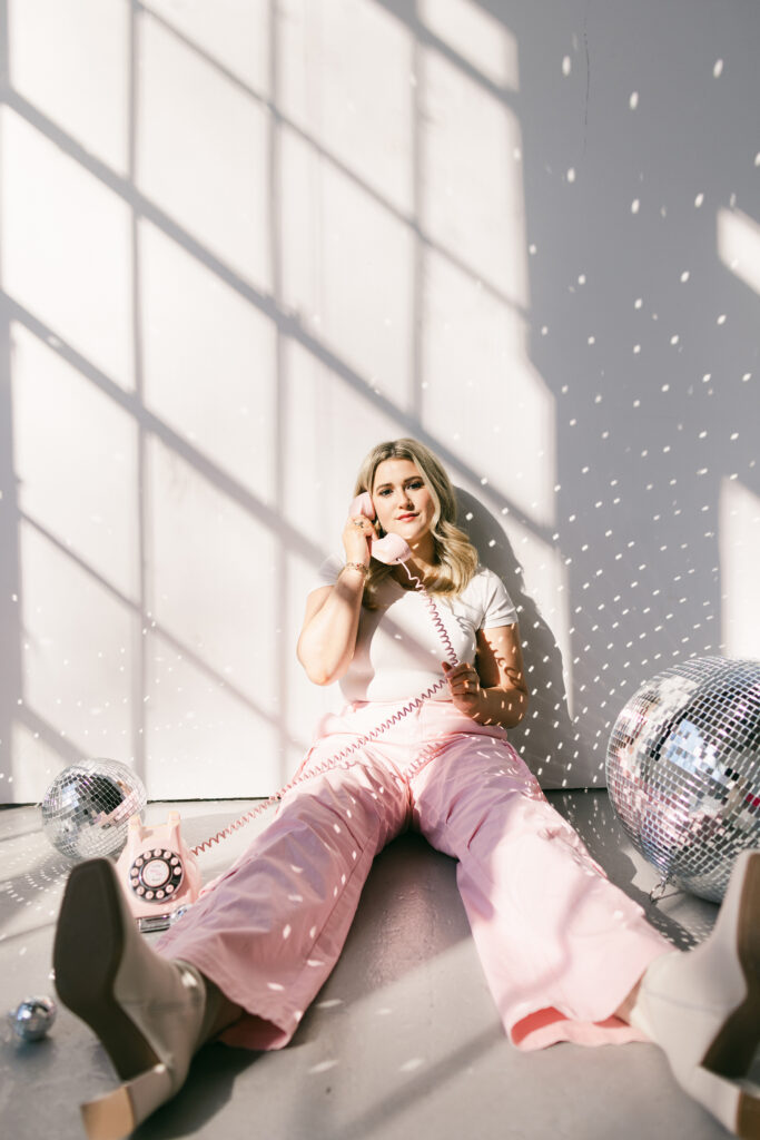 Woman sitting on studio floor in pink trousers with vintage pink phone and disco balls, surrounded by sunlight reflections – branding photography by Dani Lacey in Greenville SC.