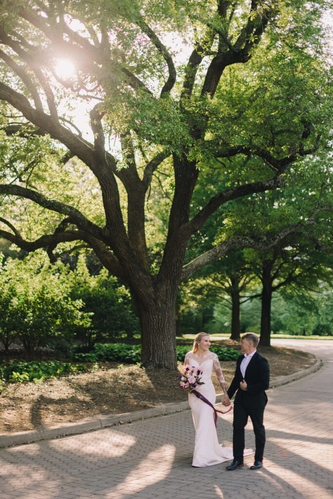 Couple walking hand-in-hand on green grass toward a lakeside view with bell tower in the distance at Furman University — one of the top easy photo spots in Greenville SC – Dani Lacey Photographs