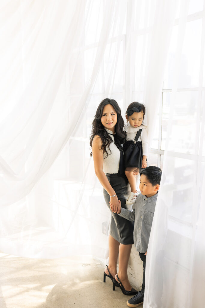 Chic mother and children in stylish black and white wardrobe posing by sheer curtains during editorial family mini session
