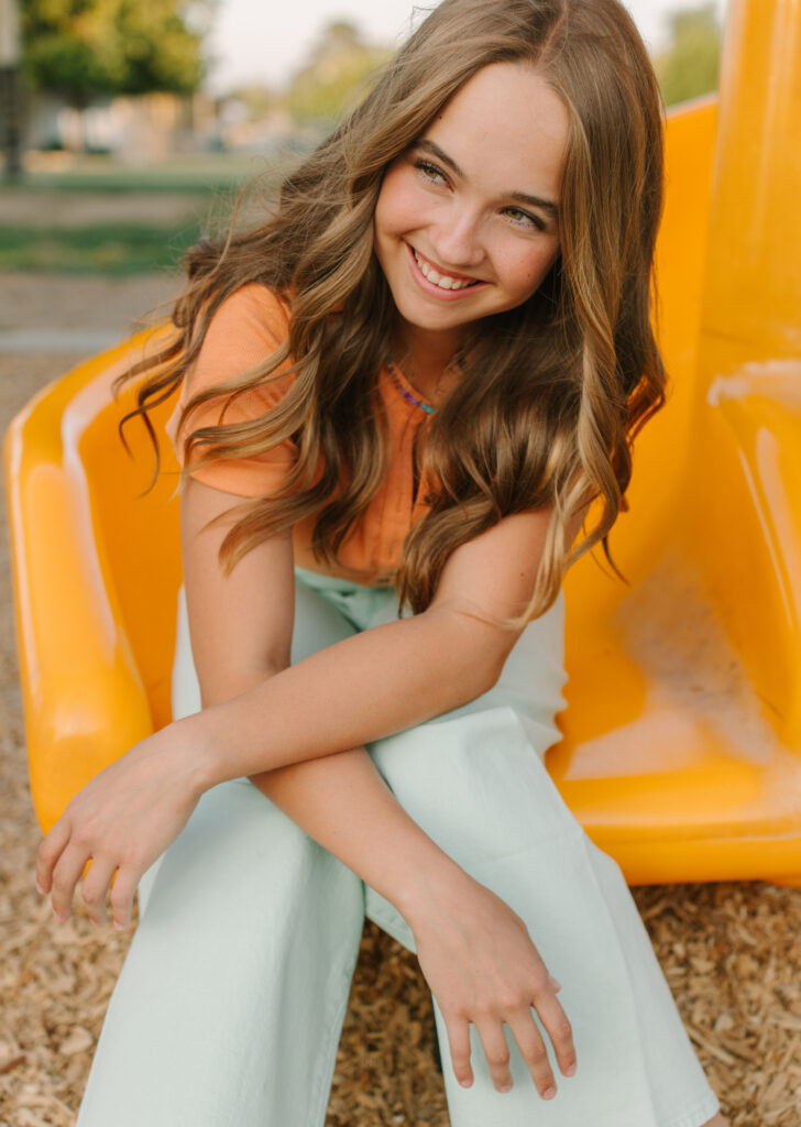 Girl with wavy hair smiles while sitting on a yellow playground slide.