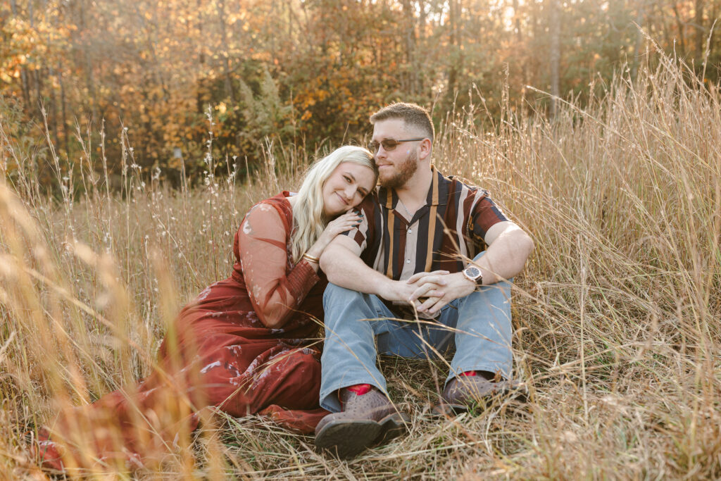 Couple sitting together in tall grass with golden trees in the background during autumn engagement photos at Campbell’s Covered Bridge. Dani Lacey Photographs.