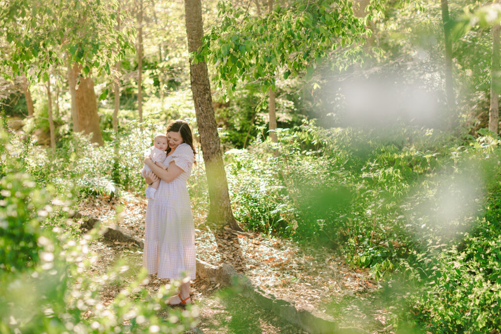 A mom stands on a wooded path holding her baby in the sunlight at Falls Park. Easy photo spots in Greenville SC