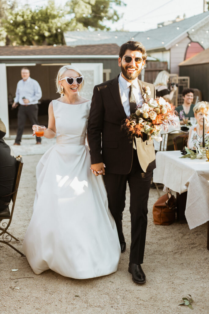 Bride and groom walk hand in hand into their backyard reception, laughing and wearing sunglasses, surrounded by golden-hour light for Building Wedding Timelines 