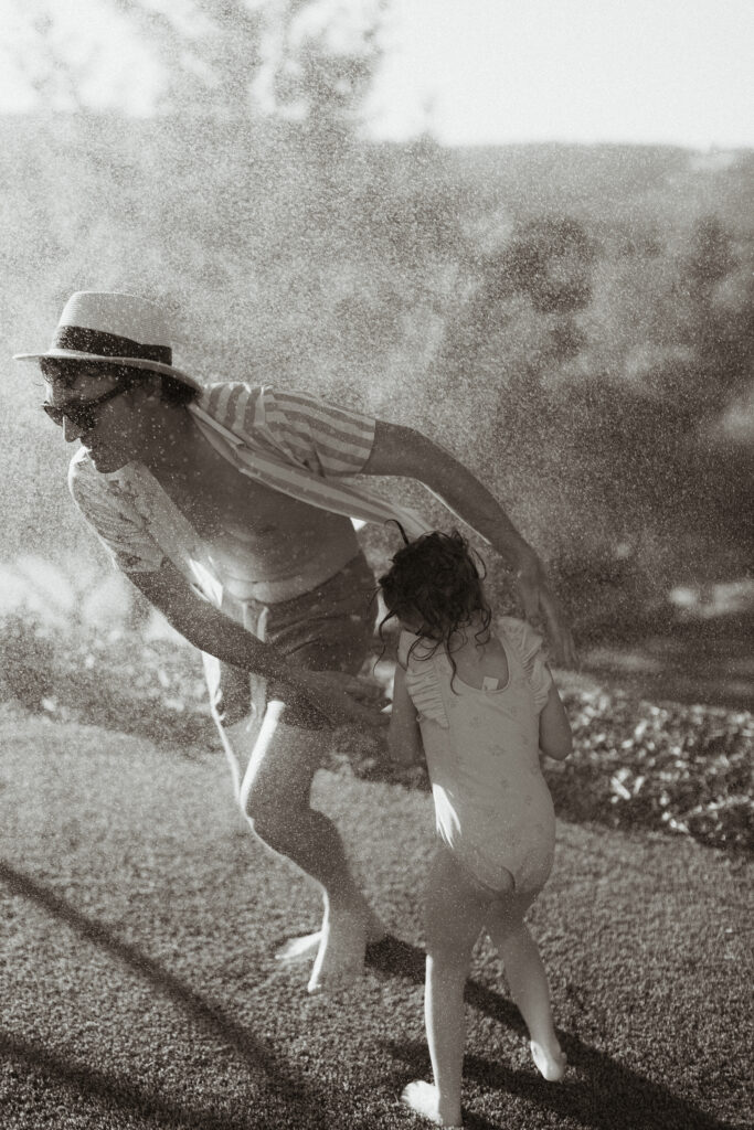 Black and white candid image of a dad and daughter running through backyard mist during a summer family shoot – Dani Lacey Photographs in Greenville South Carolina.