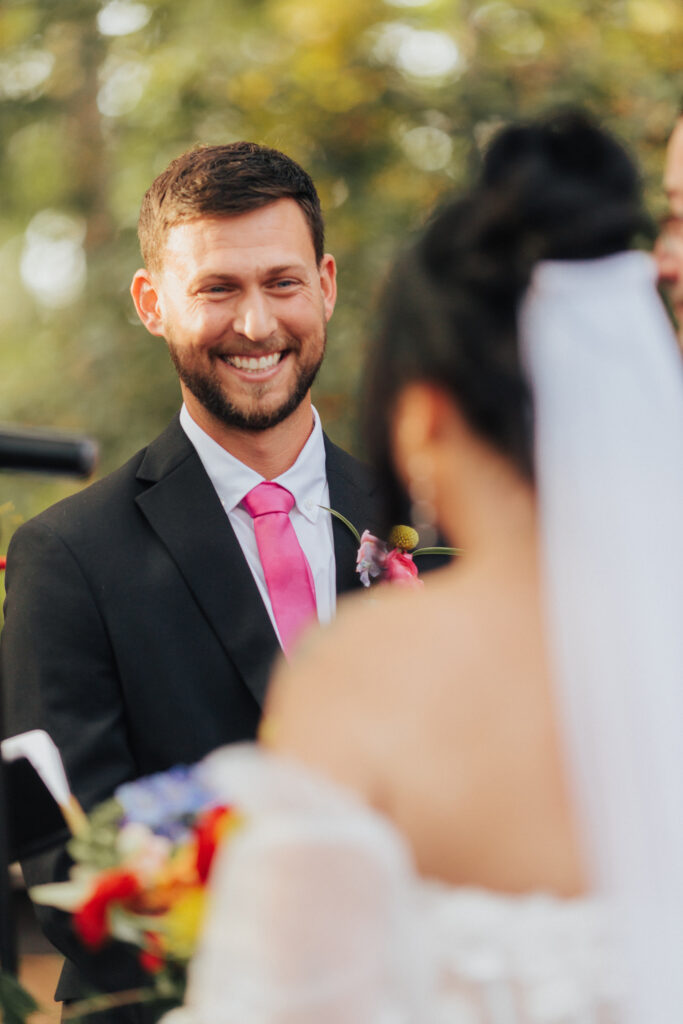 A groom in a black suit and bright pink tie smiles wide as he looks at his bride during their outdoor vows, his eyes soft with emotion.