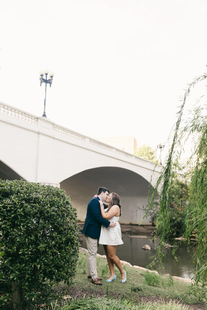 Couple kissing under a white bridge along the Reedy River during Falls Park engagement session. Dani Lacey Photographs.