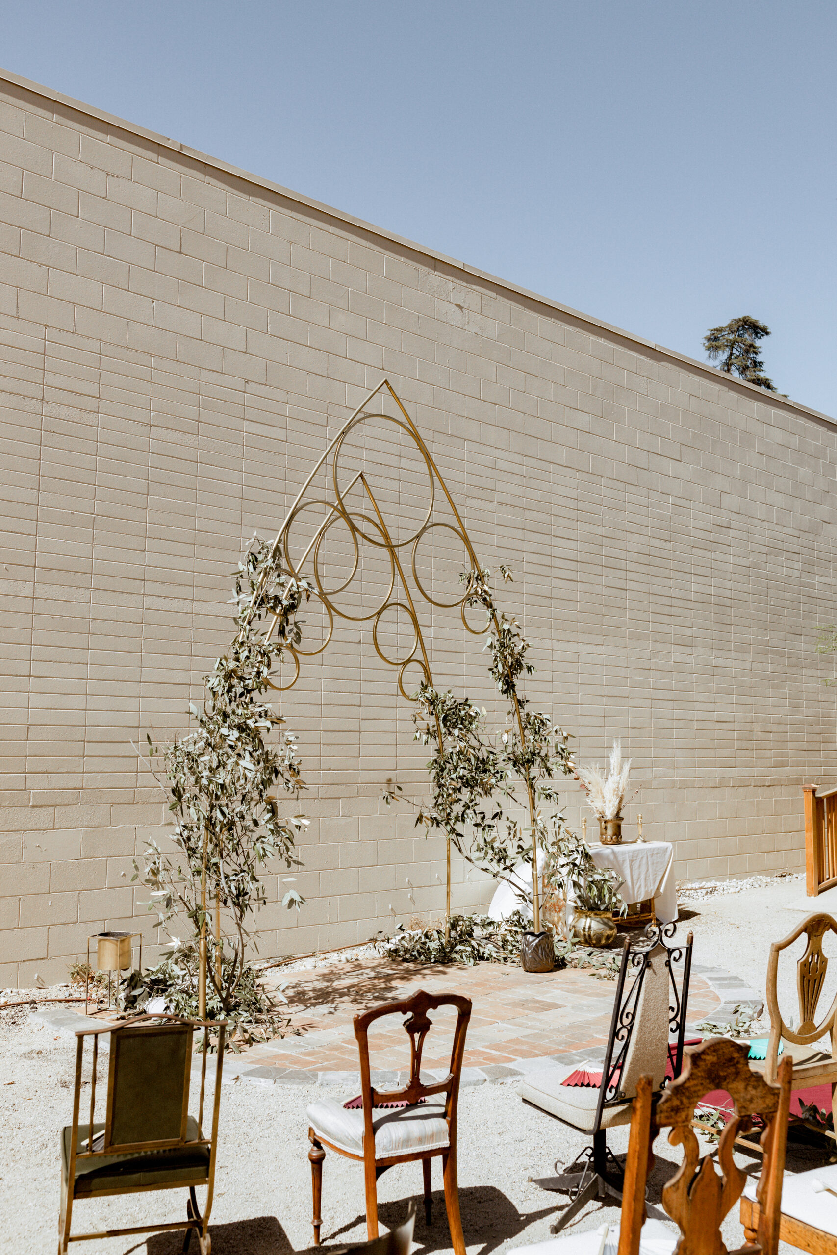 A modern gold ceremony arch with organic greenery stands against a neutral brick wall, surrounded by vintage chairs under bright daylight.