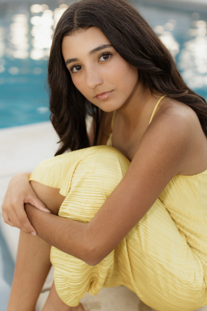 Girl in a yellow pleated dress sits beside a pool with soft lighting.
