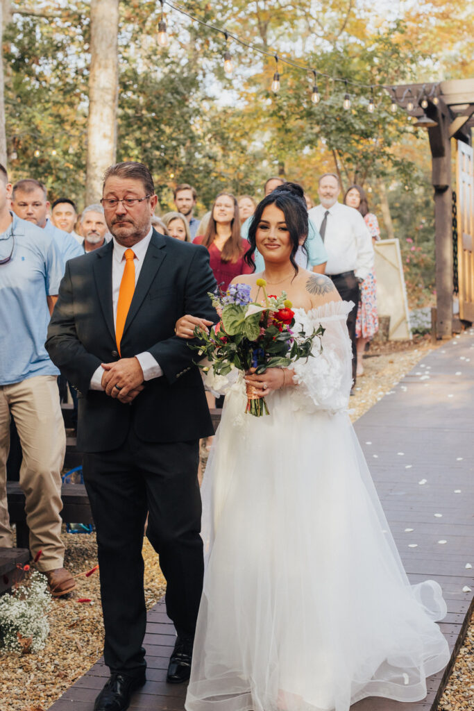 A bride with loose curls and a soft off-the-shoulder gown walks arm-in-arm with her father down an outdoor aisle, surrounded by guests watching with emotion.