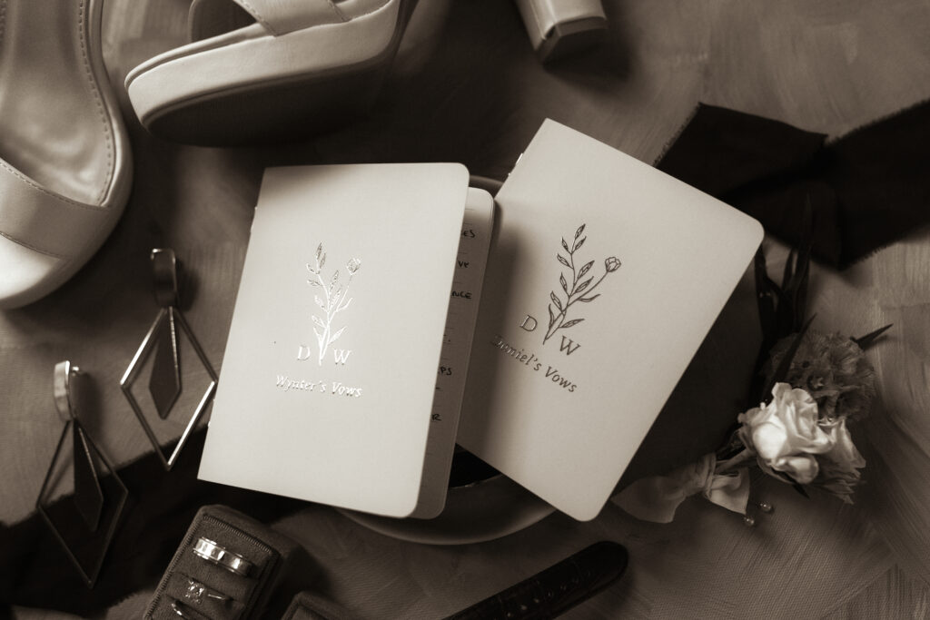 Flat lay of personalized vow books surrounded by wedding day details like earrings, shoes, rings, and a boutonnière.