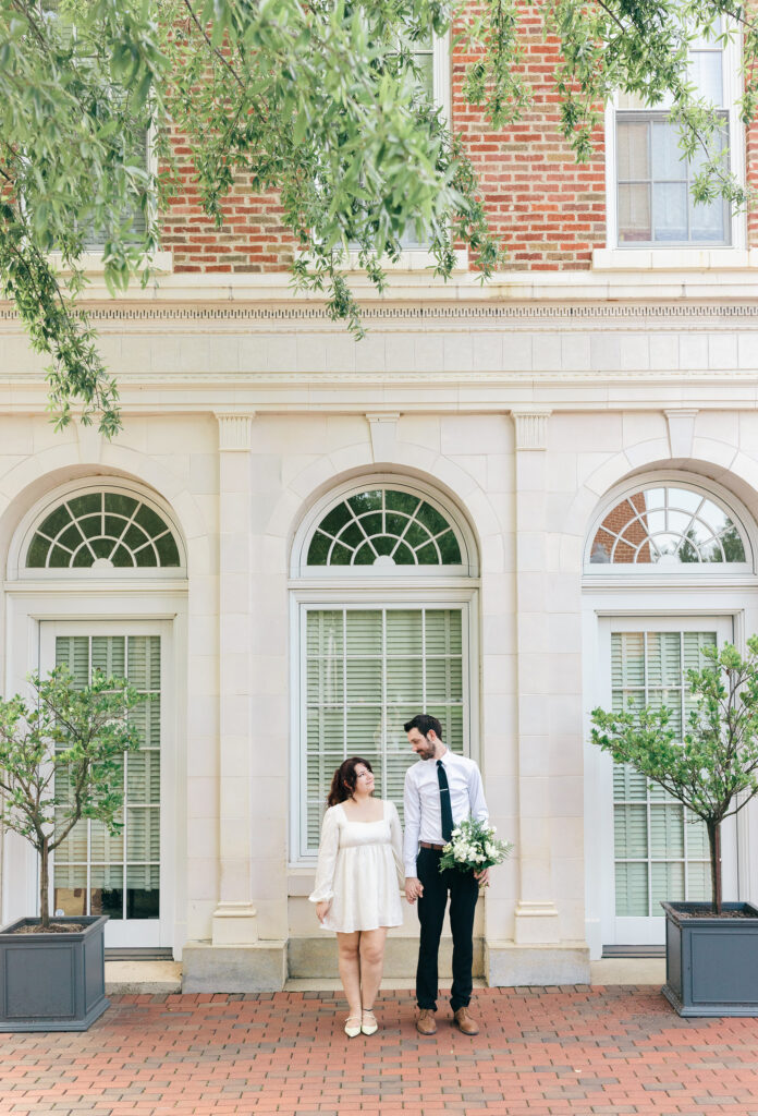 Couple holding hands in front of a historic brick building with arched windows.