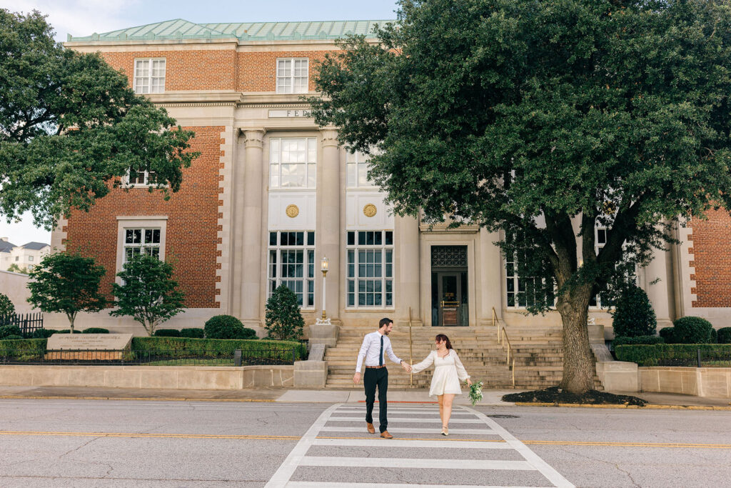 A bride and groom holding hands while crossing the street in front of the historic Spartanburg courthouse, captured by Greenville South Carolina portrait photographer Dani Lacey Photographs.