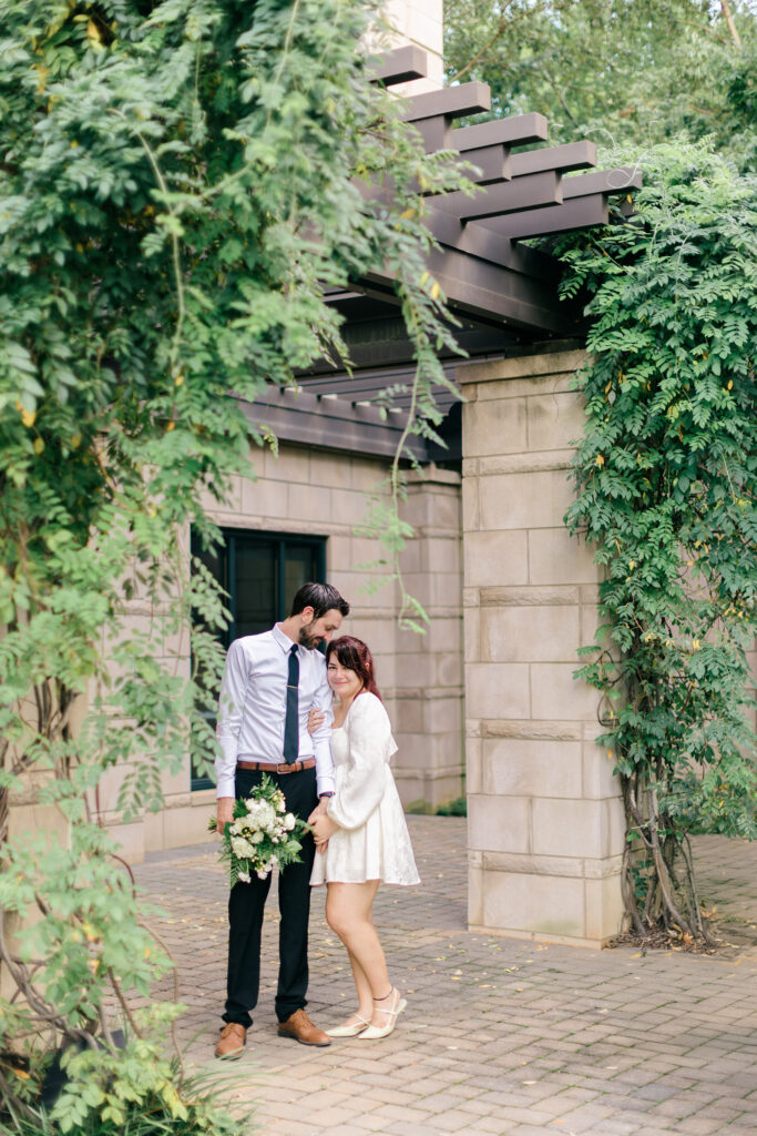 Bride and groom stand together under a vine-covered pergola in downtown Spartanburg, smiling softly at one another. Captured by Greenville SC wedding photographer Dani Lacey Photographs.
