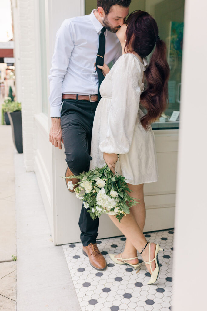 Bride and groom share a kiss outside a downtown Spartanburg storefront, with the bride holding a bouquet of white flowers. Captured by Greenville South Carolina wedding photographer Dani Lacey Photographs.