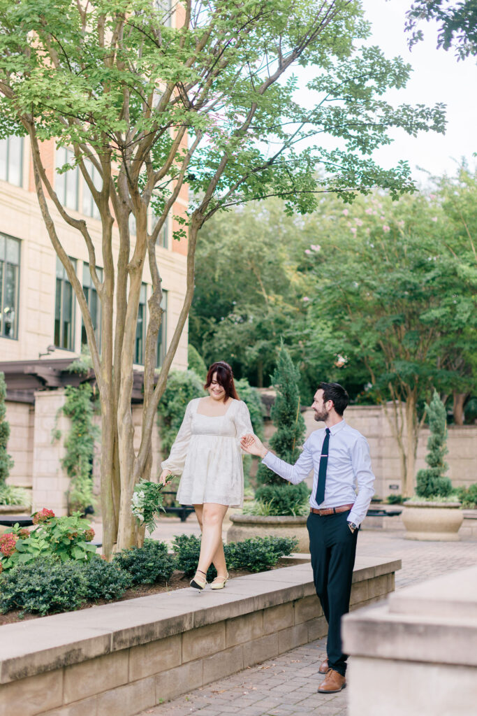 Bride in a short white dress balances on a ledge while the groom holds her hand in downtown Spartanburg, captured by Greenville South Carolina wedding photographer Dani Lacey Photographs.