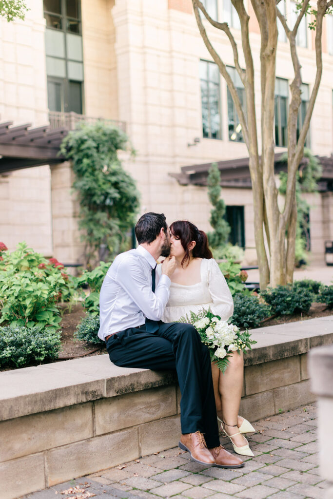 Bride and groom share a kiss while sitting on a stone ledge surrounded by greenery in downtown Spartanburg. Captured by Greenville South Carolina portrait photographer Dani Lacey Photographs.