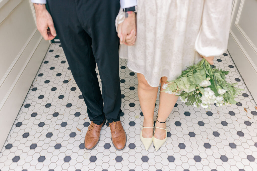 Close-up of bride and groom holding hands, showcasing their shoes and bouquet on a patterned tile floor in downtown Spartanburg. Captured by Greenville South Carolina wedding photographer Dani Lacey Photographs.