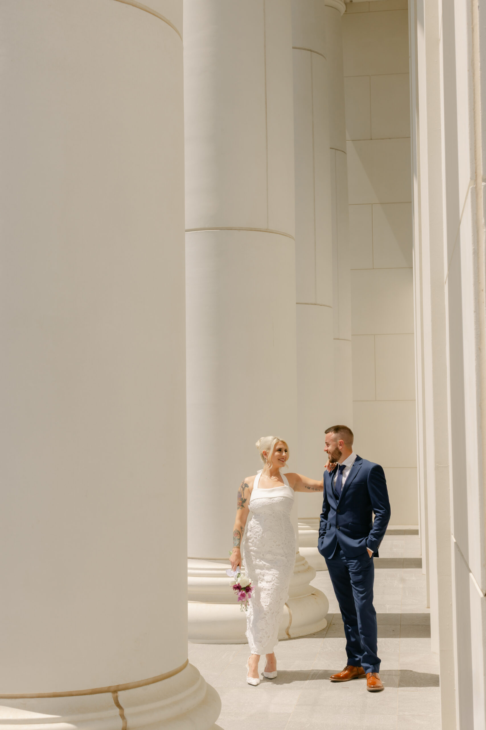 Bride and groom walk hand in hand between tall white columns at a modern courthouse in Greenville, South Carolina. Captured by Greenville SC wedding photographer Dani Lacey Photographs.