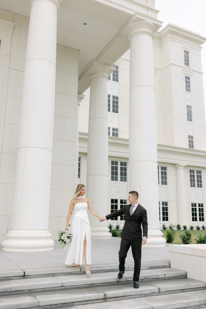 Bride and groom walk down the grand courthouse steps in Spartanburg, South Carolina, holding hands in front of tall white columns. Captured by Greenville SC wedding photographer Dani Lacey Photographs.
