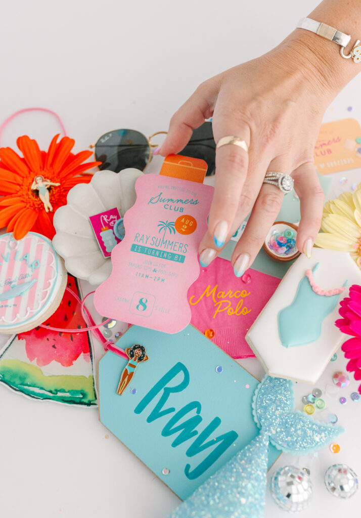 Close-up of a hand picking up a pool party invitation surrounded by themed cookies, flowers, and summer party props