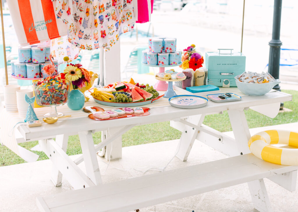 Full picnic table spread with summer fruit, cookies, flowers, snacks, and colorful details for a kids’ pool party. Dani Lacey Photographs.