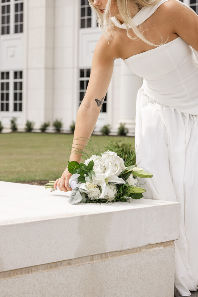 Bride in a white dress places a lush white and green bouquet on a marble ledge outside the Spartanburg County Courthouse – Spartanburg courthouse wedding photography by Dani Lacey Photographs.