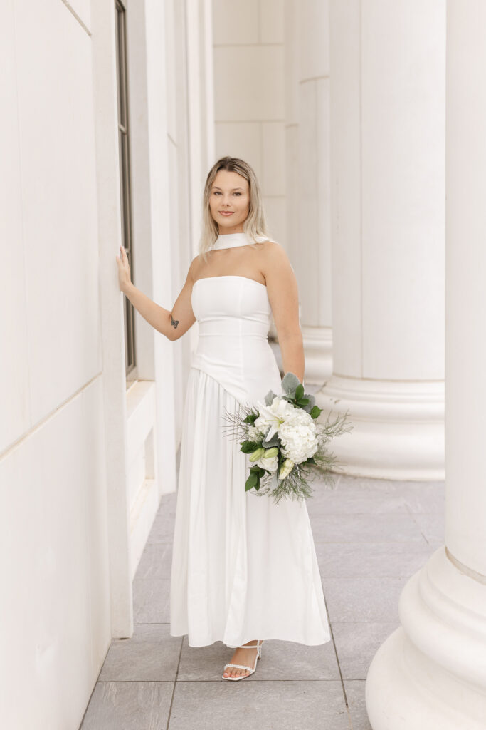 Bride in a modern white wedding gown holds a bouquet while standing among the grand white columns of the Spartanburg County Courthouse – Spartanburg courthouse wedding photography by Dani Lacey Photographs.