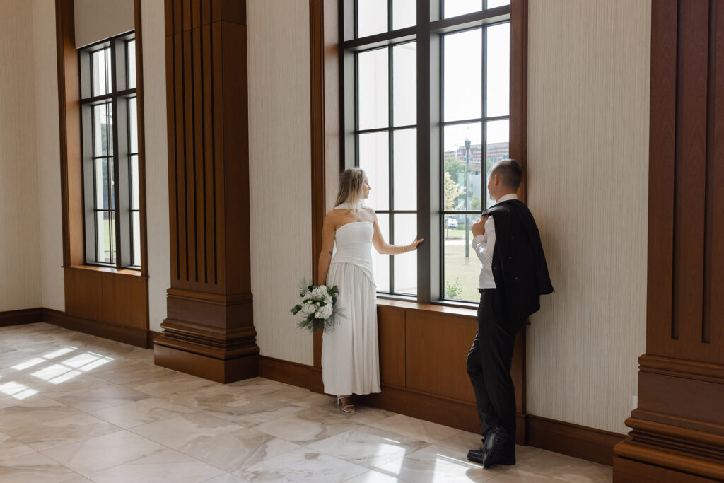 Bride in a white modern dress holding a bouquet and groom in a suit jacket over his shoulder standing by tall courthouse windows in Spartanburg, South Carolina, captured by Greenville South Carolina portrait photographer Dani Lacey Photographs.