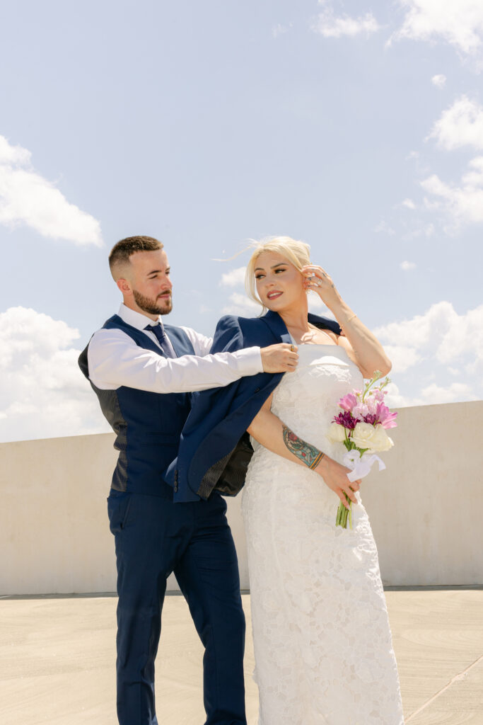 Groom helps bride into his jacket while standing outdoors at the Spartanburg Courthouse, with the bride holding a bouquet of white and purple flowers. Captured by Greenville SC wedding photographer Dani Lacey Photographs.