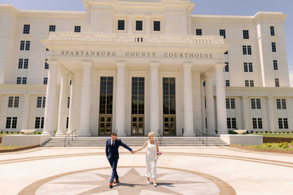 Bride and groom walk hand in hand in front of the Spartanburg County Courthouse, framed by its grand white columns. Captured by Greenville SC wedding photographer Dani Lacey Photographs.