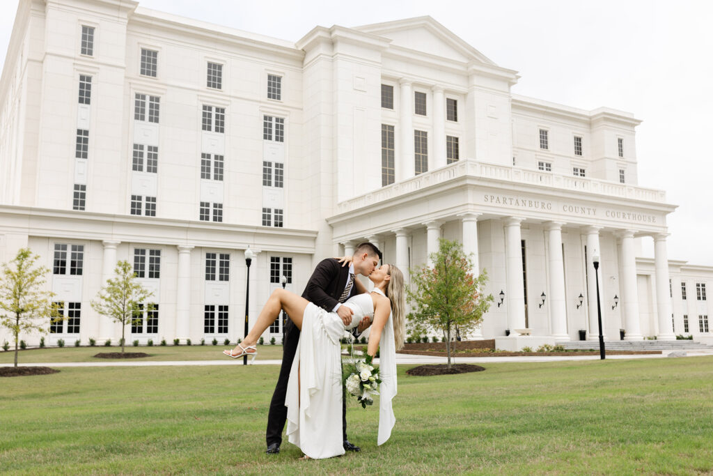 Groom in black suit dips his bride in a white wedding dress for a romantic kiss on the lawn in front of the Spartanburg County Courthouse, bouquet in hand – Spartanburg courthouse wedding photography by Dani Lacey Photographs.