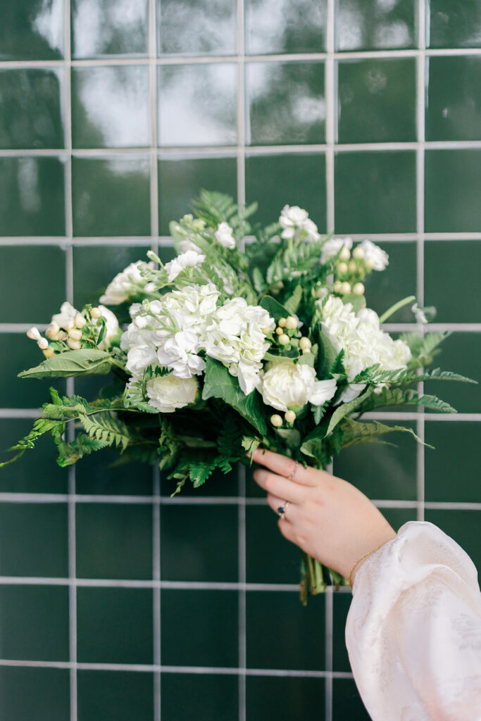 Hand holding a white and green wedding bouquet against a tiled wall.