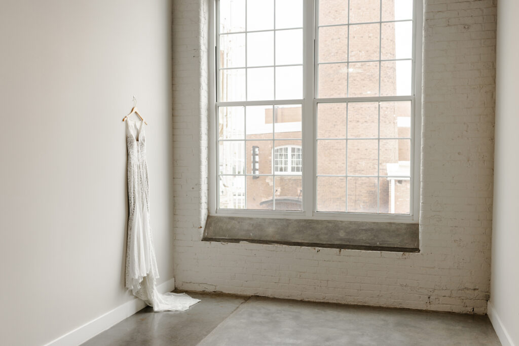 Minimalist editorial photo of wedding dress hanging on a white wall beside industrial windows at a historic favorite luxury wedding venue Judson Mill in Greenville, SC – Dani Lacey Photographs