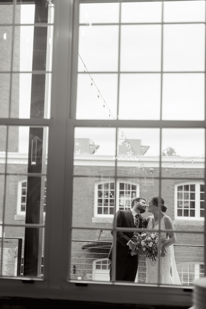 Black and white editorial wedding photo of bride and groom kissing outside industrial brick venue in Greenville, SC, captured through large window panes – Dani Lacey Photographs