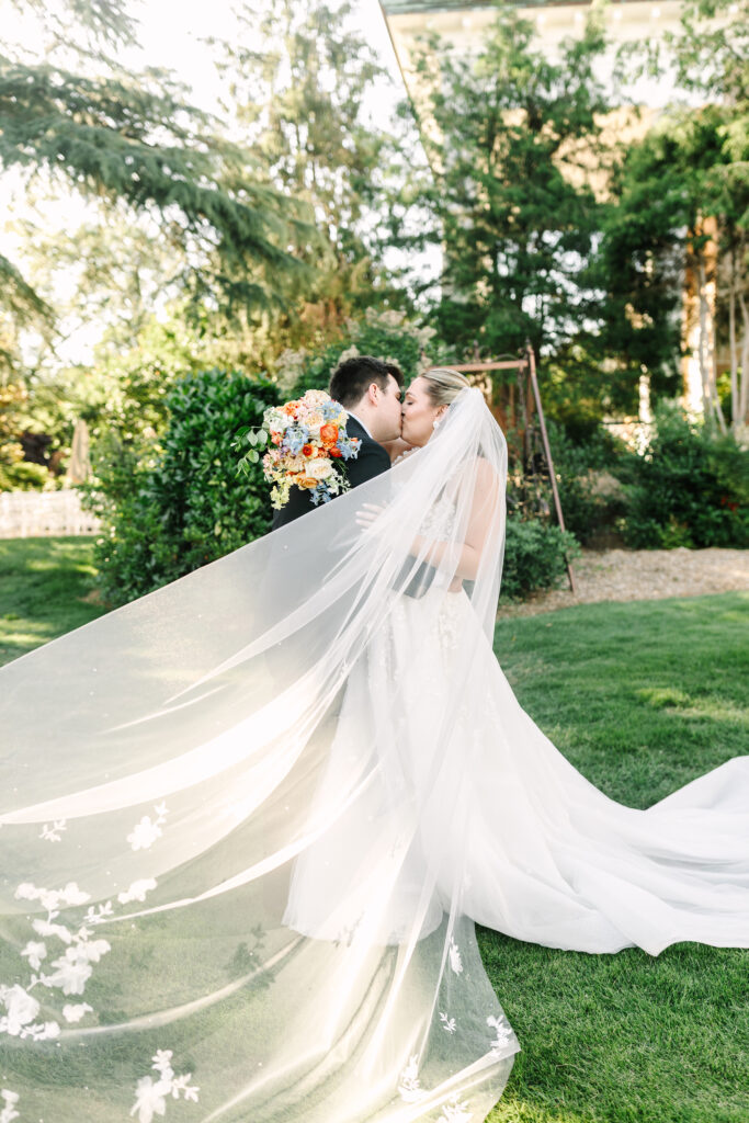 Editorial wedding photo of bride and groom kissing on the lawn at the Gassaway Mansion in Greenville, SC, with bride’s veil flowing in the wind and colorful floral bouquet – Dani Lacey Photographs